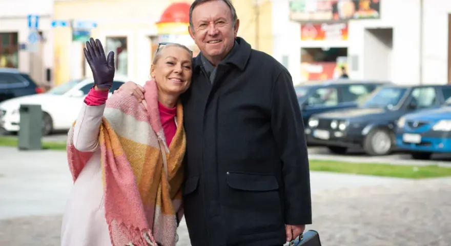 Małgorzata Szeptycka wita się z uśmiechem, obejmując partnera. W tle miejski krajobraz z samochodami.