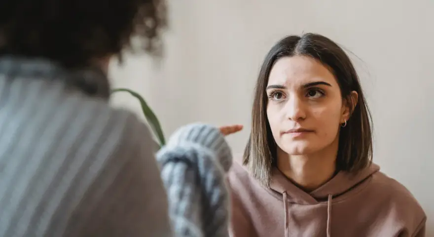 Dwie kobiety rozmawiają. Jedna wskazuje palcem na drugą, która patrzy z wyrzutem, jakby miała potworne wyrzuty sumienia.