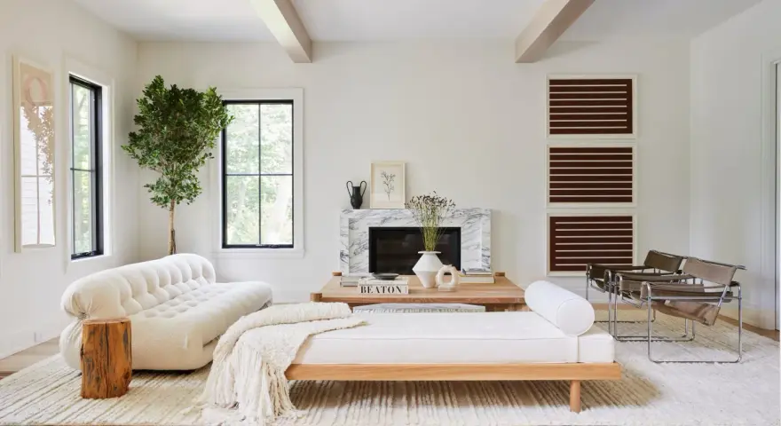 Un elegante salón blanco y madera natural, con sofá curvo, chaise longue, chimenea de mármol y sillas de cuero.