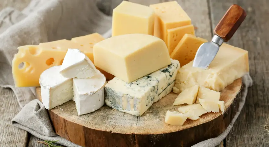 Różnorodne sery ostre gatunki: pleśniowy, żółty z dziurami, brie i parmezan, ułożone na drewnianej desce z gałązką rozmarynu.