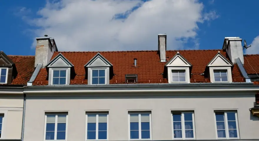 Czerwony dach z lukarnami i kominami nad jasnoszarą elewacją.