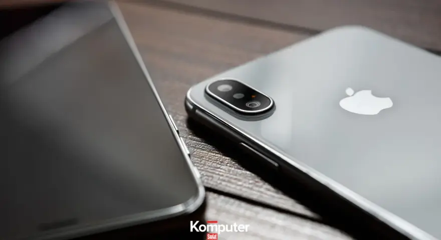 Dwa telefony leżą obok siebie na drewnianym blacie. Jeden z nich, srebrny iPhone, ma widoczny moduł aparatu. Drugi, z ciemnym ekranem, jest ustawiony bokiem. Idealne do rozmowy przez **Facetime**.
