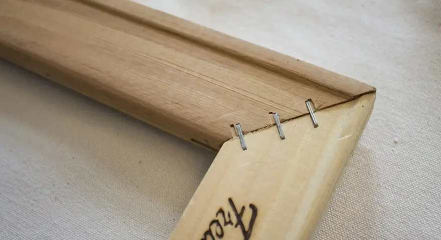 Détail d'un coin de cadre en bois, montrant comment tendre une toile sans châssis avec des agrafes.