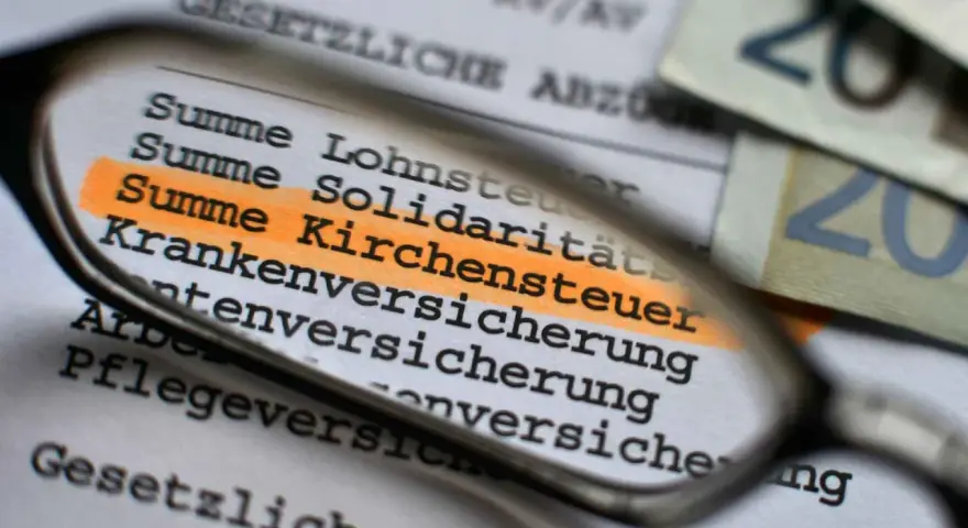 Lupe über Dokument mit hervorgehobener Kirchensteuer. Geld liegt daneben.