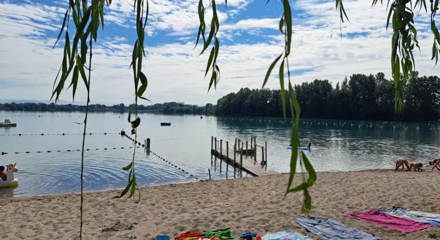 Ein sonniger Tag am Lauterbourg See. Handtücher liegen am Sandstrand, Kinder planschen im Wasser.