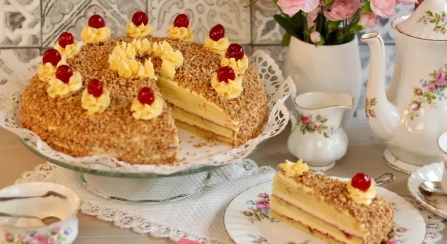 Ein köstlicher Frankfurter Kranz, verziert mit Buttercreme-Tupfern und Kirschen, umgeben von gehackten Nüssen. Ein Stück ist auf einem Teller angerichtet.