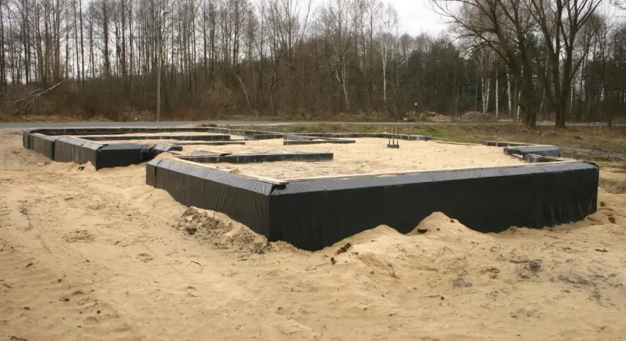 Gotowe ławy fundamentowe otoczone czarną folią, czekają na zalanie betonem. W tle las.