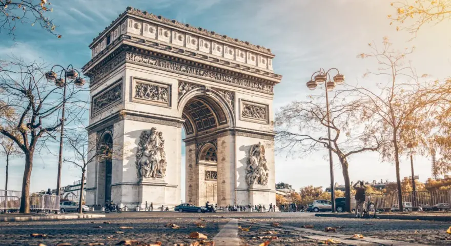 La majestuosa entrada del Arco del Triunfo en París, con esculturas y detalles ornamentales, bajo un cielo azul y árboles otoñales.