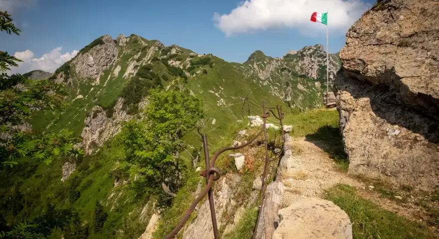 Sentiero della Pace tra montagne verdi, con bandiera italiana che sventola. Un percorso che ricorda la storia.