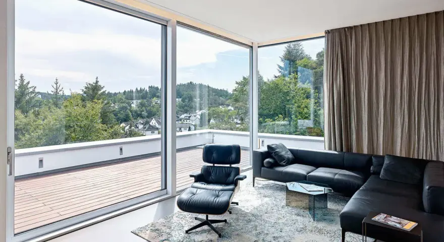 Nowoczesny salon z panoramicznym oknem narożnym, czarną skórzaną kanapą i fotelem Eames. Widok na zielone wzgórza.
