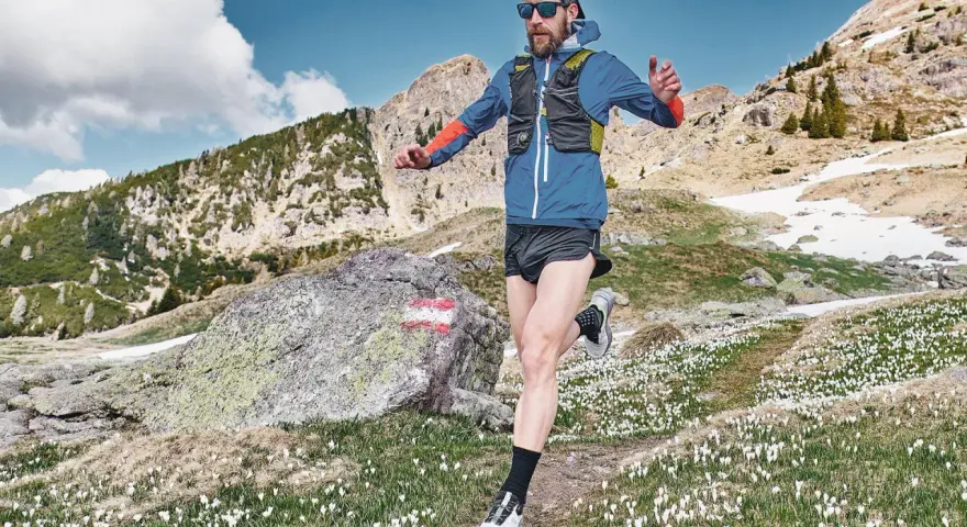 Uomo con barba corre in montagna, circondato da crochi bianchi e rocce. Il sentiero si snoda tra prati e pendii innevati.