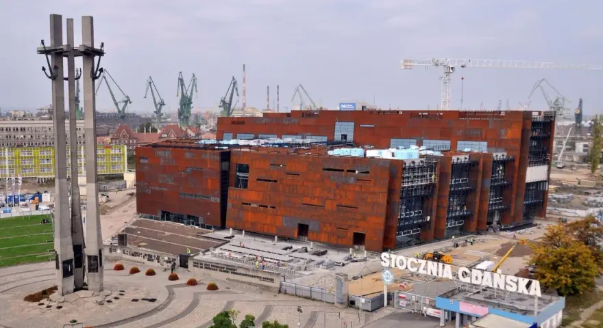 Nowoczesne muzeum na terenie Stoczni Gdańskiej, z charakterystycznymi, rdzawymi fasadami, w otoczeniu dźwigów stoczniowych i Pomnika Poległych Stoczniowców.