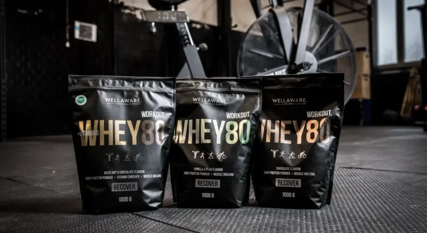 Tre påsar med Wellaware Whey80 proteinpulver står uppradade på ett gymgolv. Perfekt för återhämtning efter träning, oavsett om du föredrar vassle eller kasein.