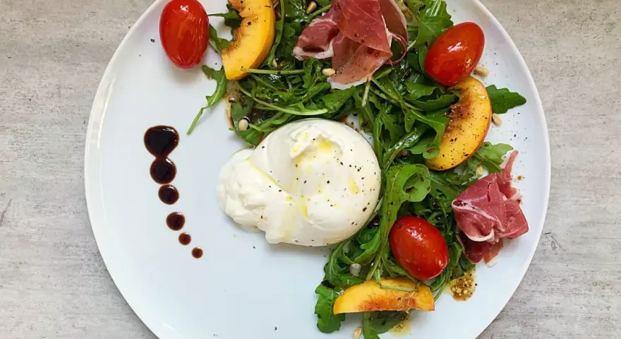 Burrata mit Rucola, Tomaten, Pfirsichspalten und Prosciutto auf einem Teller.