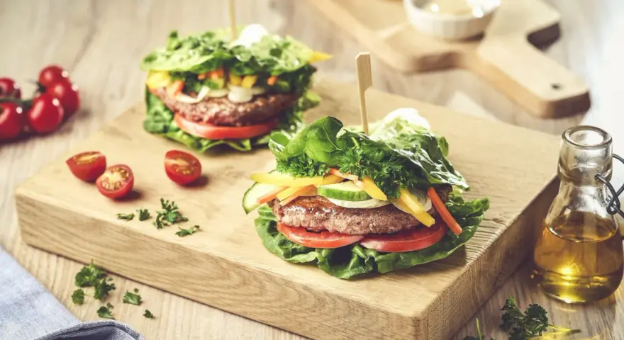 Dwa soczyste burgery w sałacie, z pomidorami, ogórkiem i papryką, na drewnianej desce.