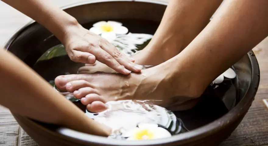 Soaking feet in a warm bath with Epsom salt for feet infection. A hand gently massages the foot, surrounded by water and white flowers.