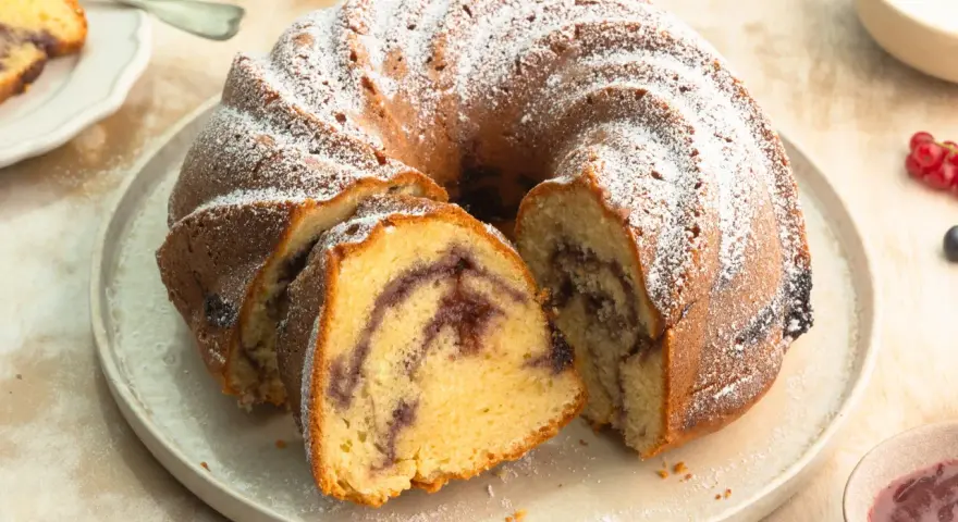 Ein Gugelhupf mit Marmelade, bestäubt mit Puderzucker. Ein Stück ist bereits angeschnitten und zeigt die köstliche Füllung.
