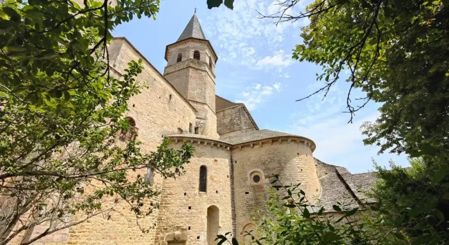 Vue d'une église romane à Villeneuve de Rouergue, encadrée par des arbres verdoyants sous un ciel bleu parsemé de nuages.