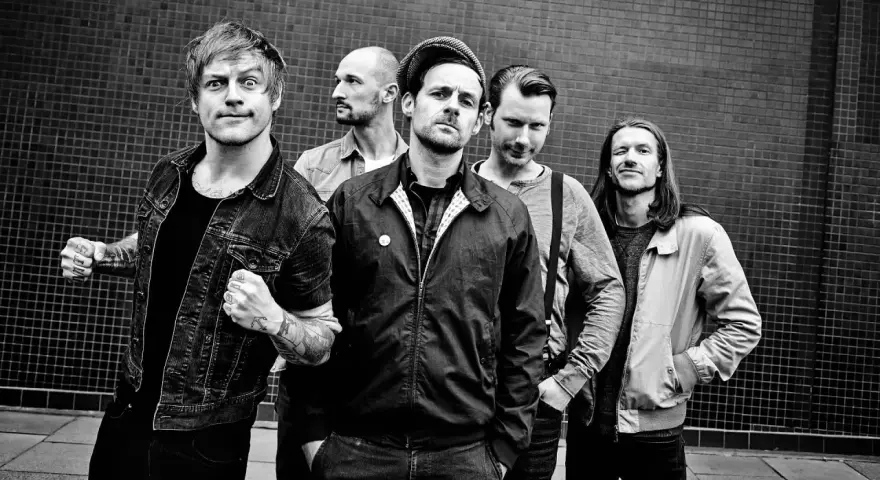Fünf Männer stehen vor einer gemusterten Wand. Sie sehen aus wie eine energiegeladene Punkband, bereit, die Bühne zu rocken.