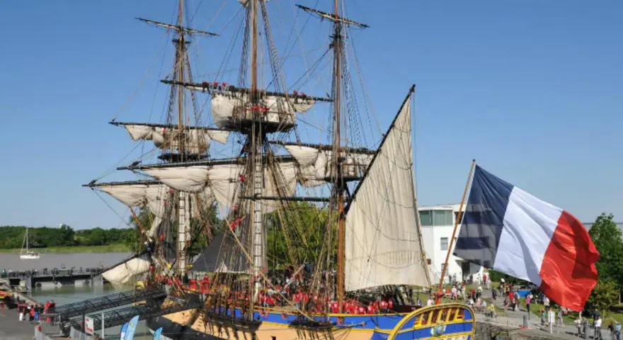 Le majestueux bateau l'Hermione, réplique d'une frégate du XVIIIe siècle, est amarré, pavoisé du drapeau français. Une foule s'est rassemblée pour admirer ce navire historique.