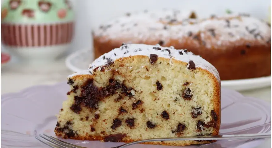 Una fetta di soffice torta con gocce di cioccolato, cosparsa di zucchero a velo, su un piatto lilla con una forchetta.