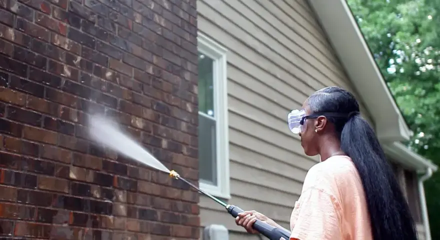 Woman pressure washing a dark acid wash brick wall, restoring its natural beauty.