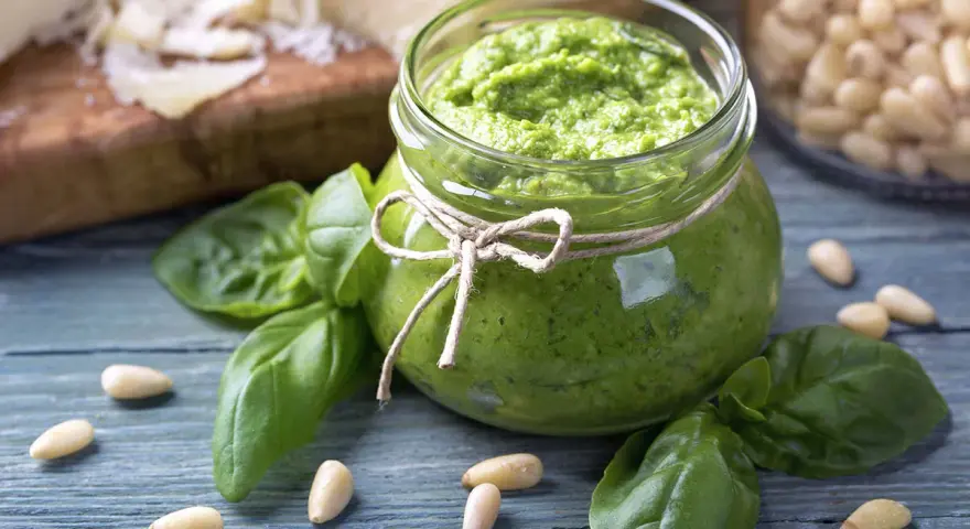 Słoik domowego pesto z bazylii, otoczony świeżymi liśćmi bazylii i orzeszkami pinii. Obok kawałki parmezanu. Idealne do makaronu.