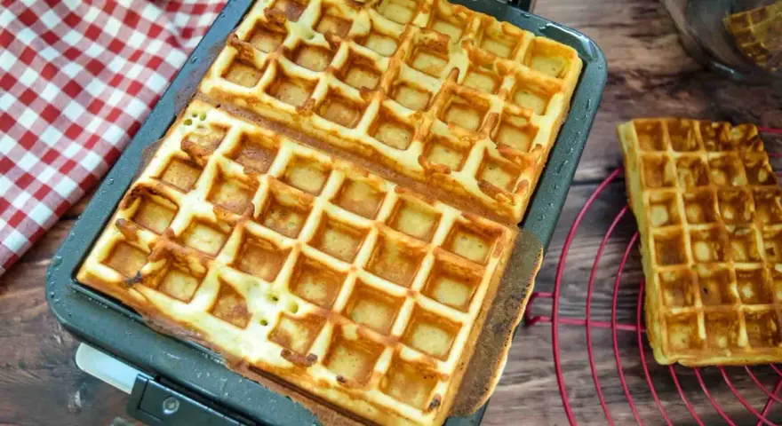 Frisch gebackene belgische Waffeln in einer Waffelform, daneben eine auf einem Rost.
