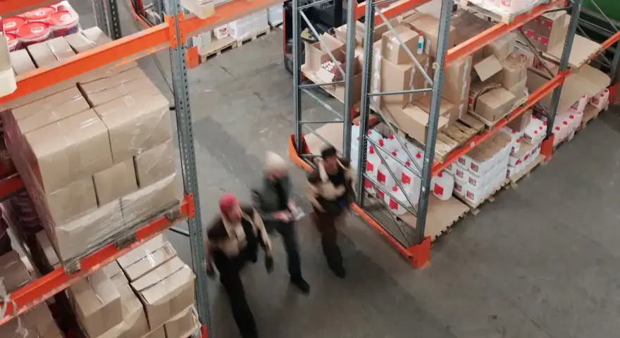 Magazzino con scaffalature piene di scatoloni e taniche. Tre persone camminano velocemente, forse per svolgere mansioni di aiuto magazziniere.