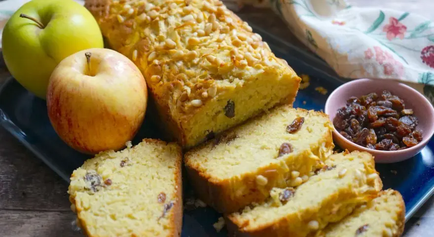 Plumcake alle mele dorato, con pinoli e uvetta, accompagnato da mele fresche e una ciotolina di uvetta.