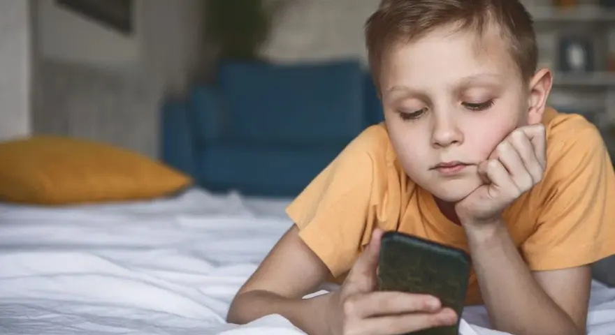 Chłopiec leży na łóżku, patrząc na telefon. Zastanawia się, co to jest sexting i jakie niesie zagrożenia.