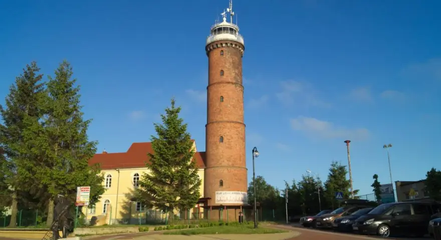 Wysoka, ceglana latarnia morska Jarosławiec góruje nad placem z drzewami i budynkiem.