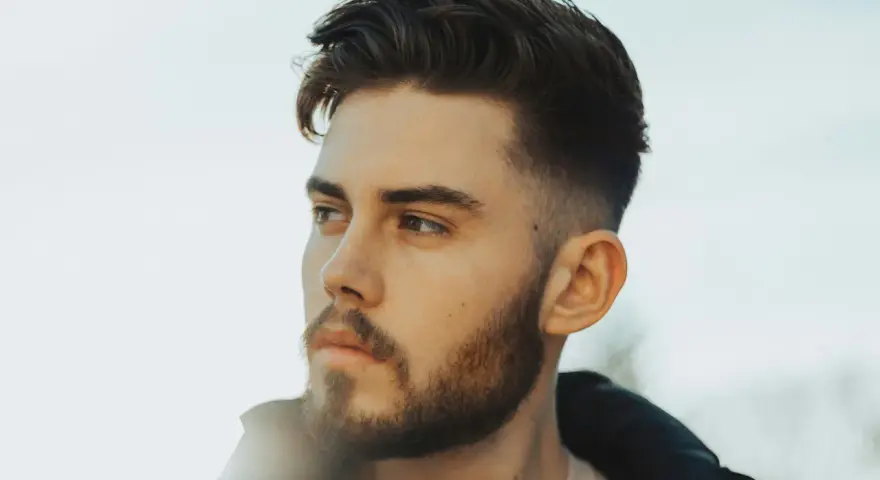 Ein Mann mit Bart und einem modernen taper fade design im Haar blickt nachdenklich zur Seite.