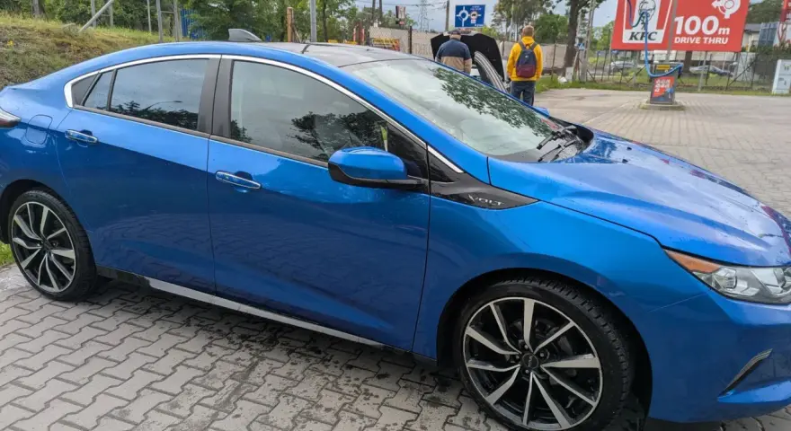 Błękitny Chevrolet Volt z czarnym dachem, zaparkowany na mokrej kostce brukowej. W tle widać stację KFC.