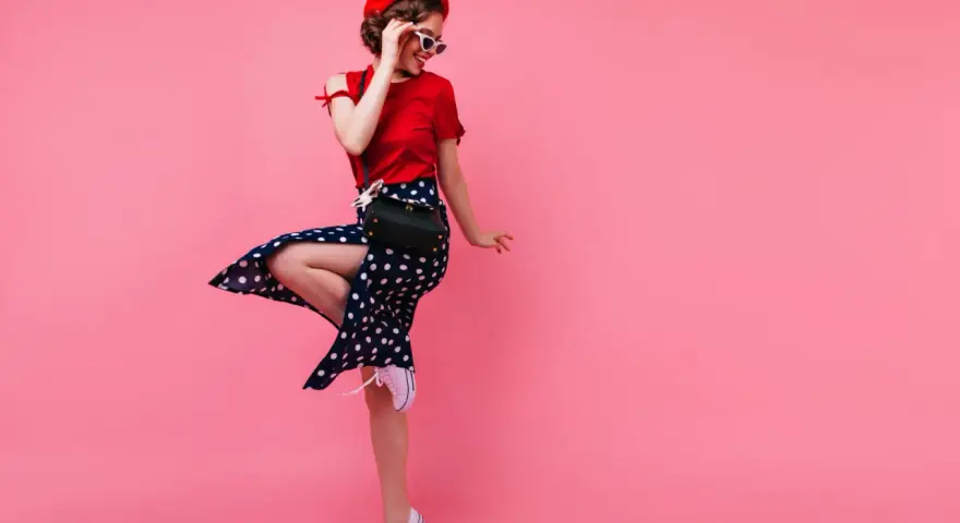 Dziewczyna w czerwonym beretce i sukience w grochy, w stylu pin up, skacze na różowym tle.