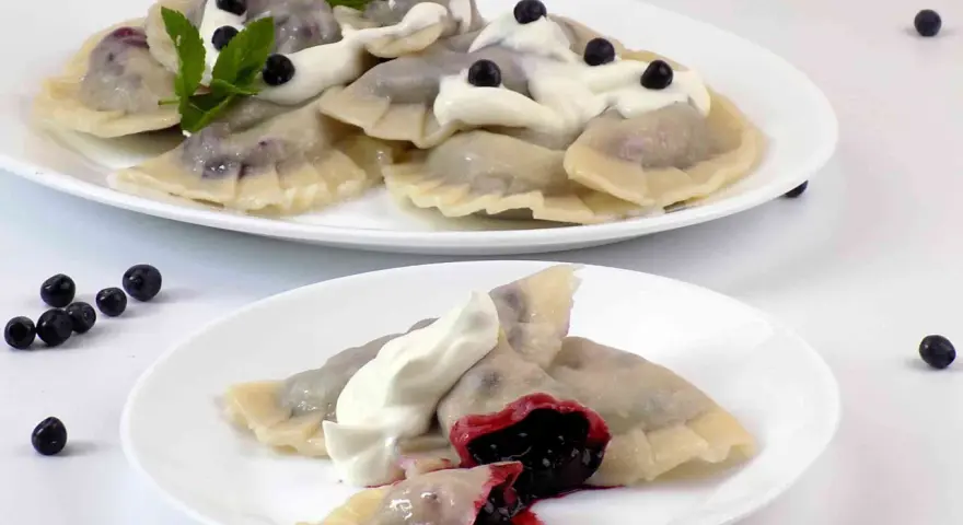 Pyszne pierogi z jagodami, polane śmietaną i ozdobione miętą. Na talerzu widać też kilka świeżych jagód.