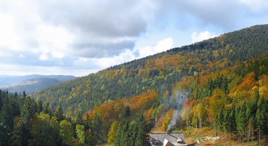 Jesienny krajobraz w niskich górach w Polsce. Drewniany dom z dymem z komina, otoczony lasem w barwach jesieni.