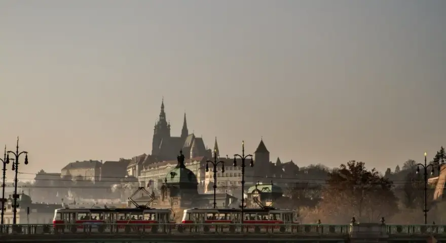 Most w Czechach, po którym przejeżdża tramwaj, z widokiem na zabytkowe budowle w oddali.