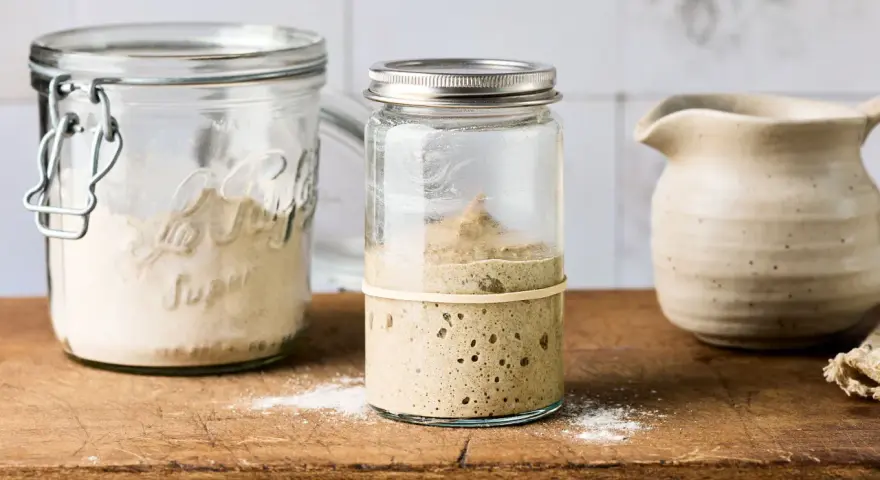 Blubbernder Sauerteig im Glas, bereit für schnelles Sauerteig herstellen. Daneben ein Glas mit Mehl und ein Krug.