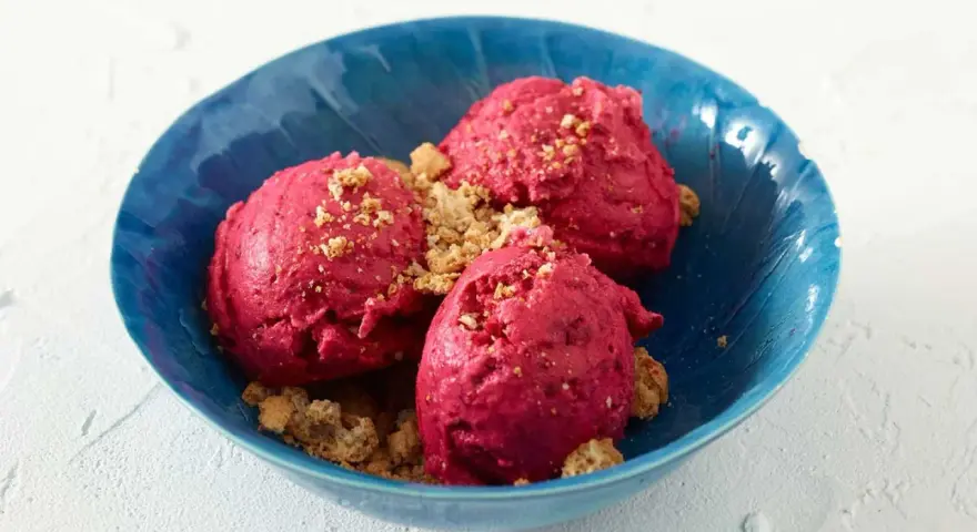 Drei Kugeln leuchtend pinkes Sauerrahm Eis, bestreut mit knusprigen Krümeln, in einer blauen Schale.