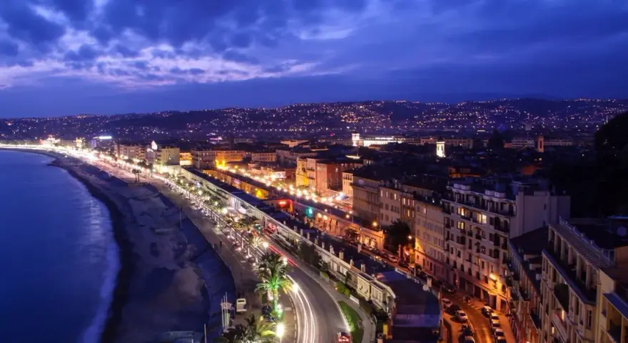 Night view of the Promenade des Anglais from Castle Hill, Nice, France, with city lights twinkling and car trails on the road.