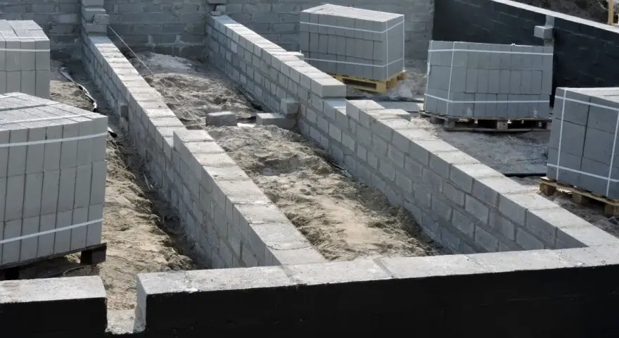 Fundamenty z bloczków betonowych, przygotowane do prac związanych z hydroizolacją. Na placu budowy widać też palety z materiałami.
