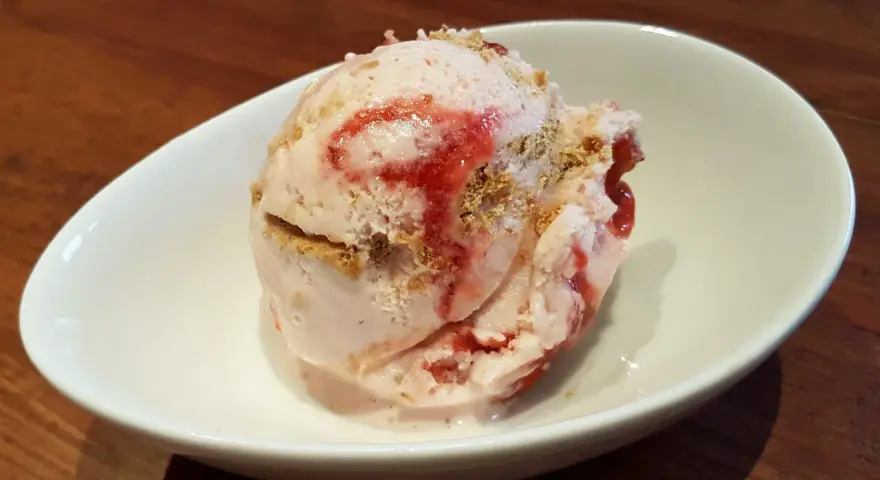 Ein Löffel cremiges strawberry cheesecake eis mit Erdbeersauce und Keksstückchen in einer weißen Schale.