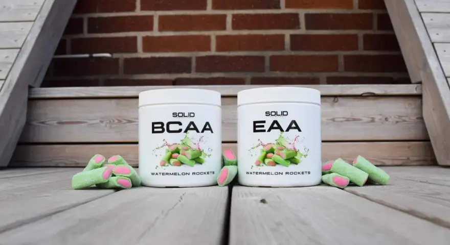 Två burkar med kosttillskott, en märkt BCAA och en EAA, står framför en tegelvägg. Runt burkarna ligger gröna godisar med rosa inuti, som ser ut som vattenmelonraketer.