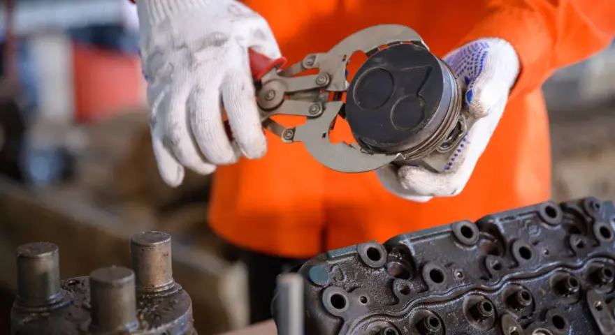 Mechanik w białych rękawicach trzyma tłok z widocznymi objawami zużytych pierścieni tłokowych. Obok leży głowica silnika.