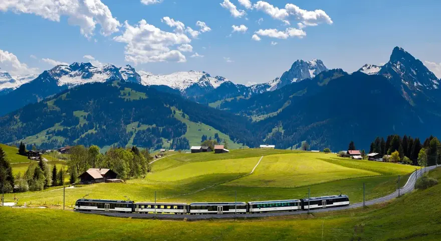 A train travels through a lush green valley with snow-capped mountains in the background. Many positive goldenpass line reviews mention this scenic route.
