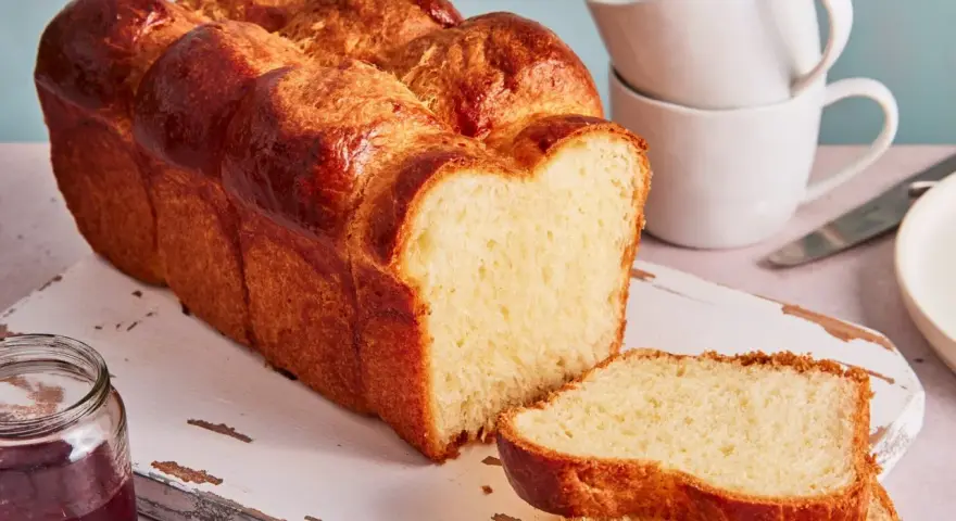 Ein goldbraunes Brioche Brot Rezept, aufgeschnitten auf einem Holzbrett mit Marmelade und Tassen im Hintergrund.