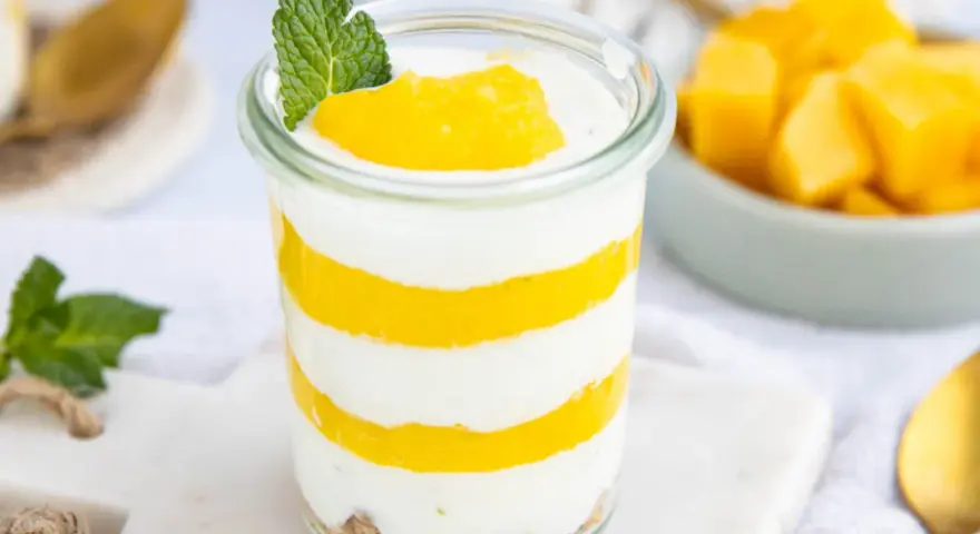 Schichtweise geschichtetes Mango-Parfait in einem Glas, garniert mit Minze. Daneben eine Schüssel mit frischen Mangostücken.