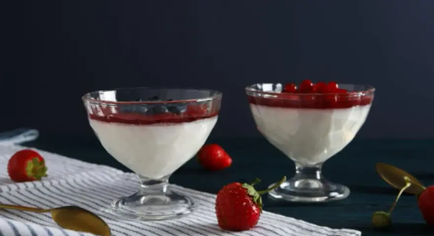 Dwie panna cotta: jedna na śmietance z borówkami, druga na jogurcie z malinami. Obok świeże truskawki i złote łyżeczki.