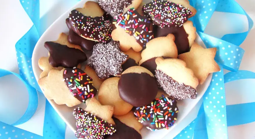 Un plato lleno de pastas de té caseras, algunas cubiertas de chocolate y chispas de colores, otras con coco rallado, rodeadas de cintas azules.